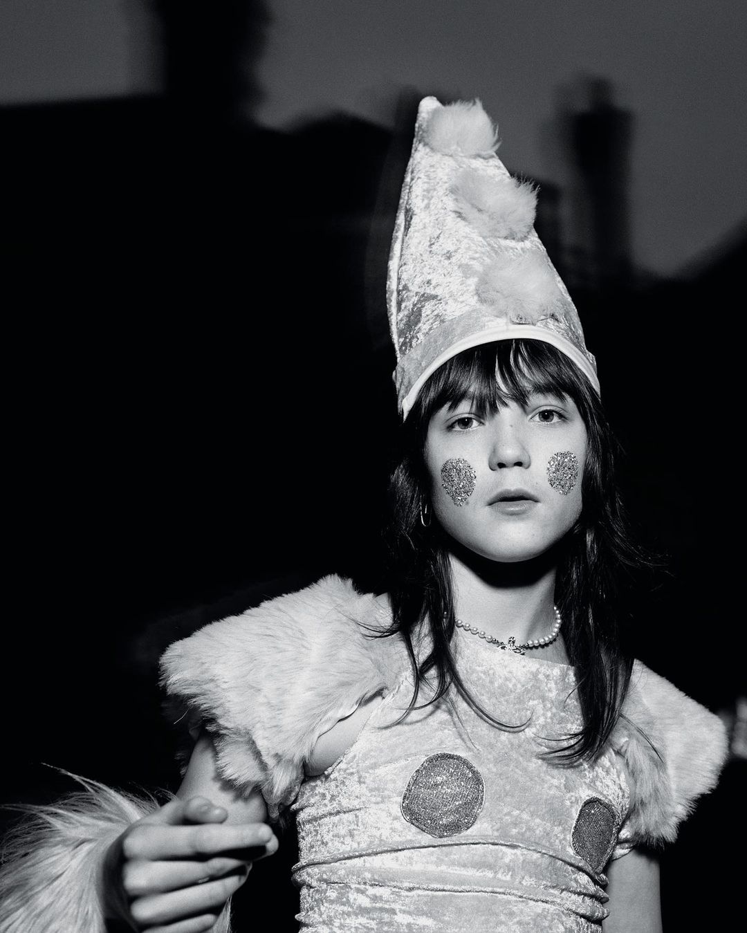 Black and white portrait of a child wearing a sparkly costume and pointy hat with pom poms, adorned with glittery face makeup.