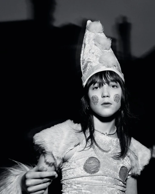 Black and white portrait of a child wearing a sparkly costume and pointy hat with pom poms, adorned with glittery face makeup.