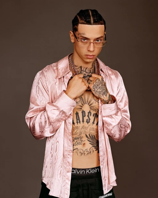 Portrait of a young adult man with braids, tattoos, glasses, and jewelry wearing a pink satin shirt against a brown background.