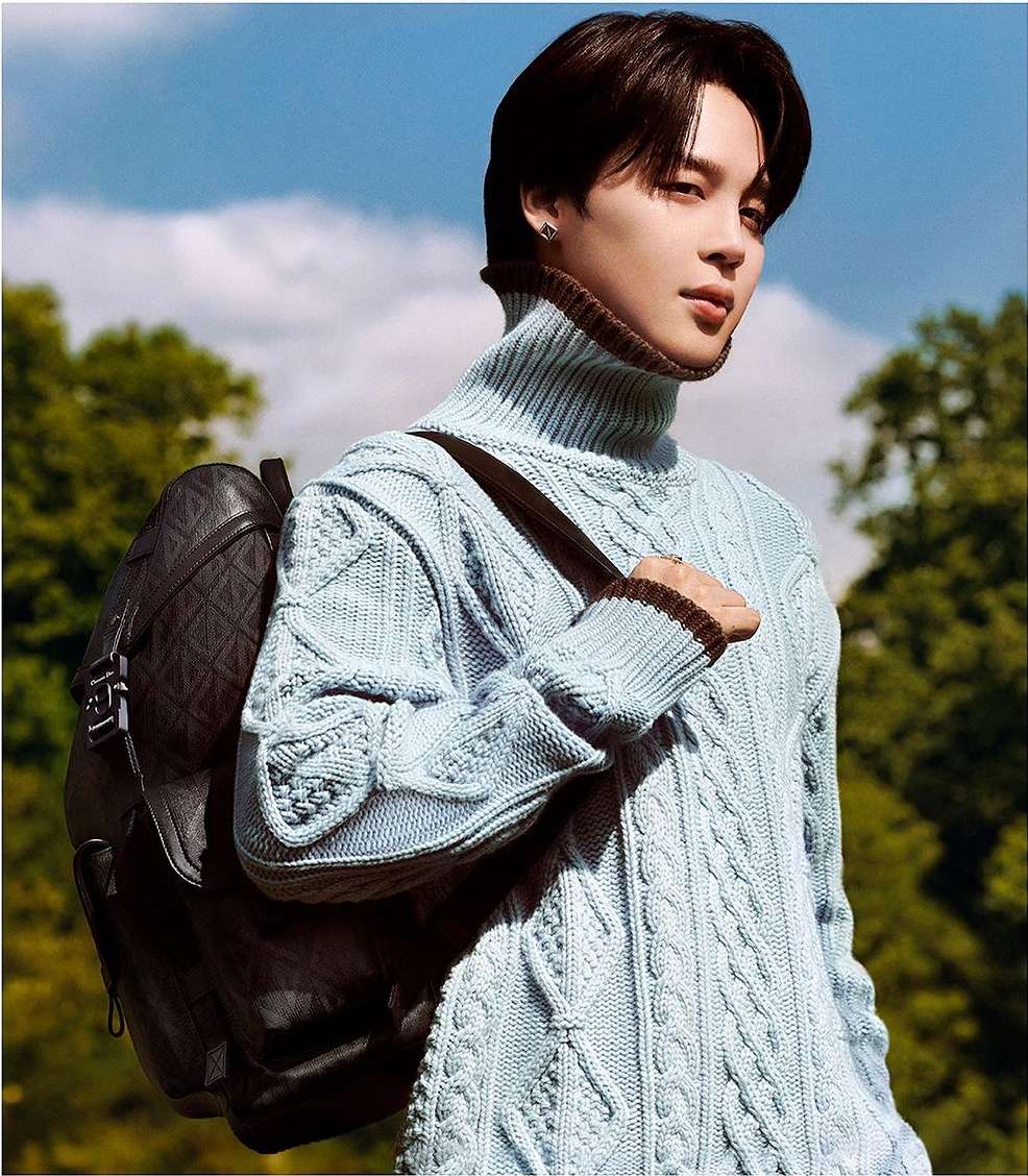 Adult male wearing a light blue turtleneck sweater poses with a black backpack against a background of trees and blue sky.