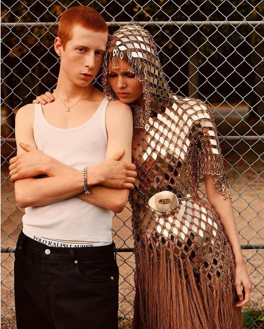 Two young adult models pose together outdoors against a chain-link fence; one is a man in a white tank top and the other wears a silver, fringed dress.