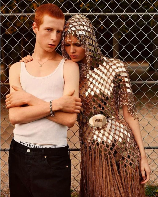 Two young adult models pose together outdoors against a chain-link fence; one is a man in a white tank top and the other wears a silver, fringed dress.
