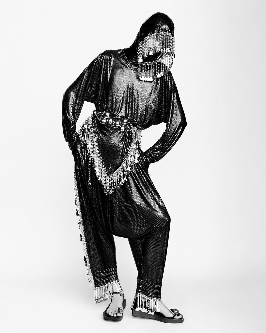 Person in a black chainmail-like outfit with a face covering and tassels posing against a white backdrop in a studio setting.
