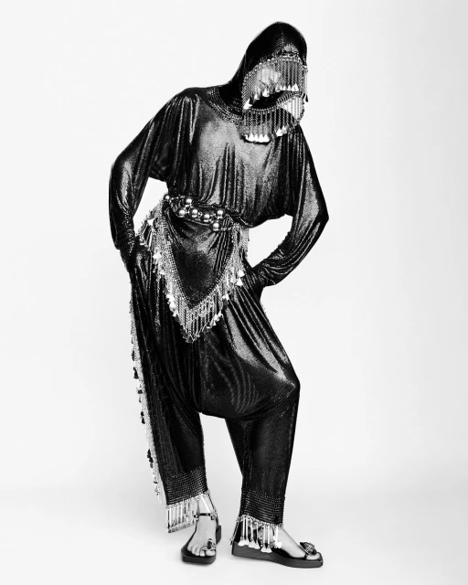 Person in a black chainmail-like outfit with a face covering and tassels posing against a white backdrop in a studio setting.