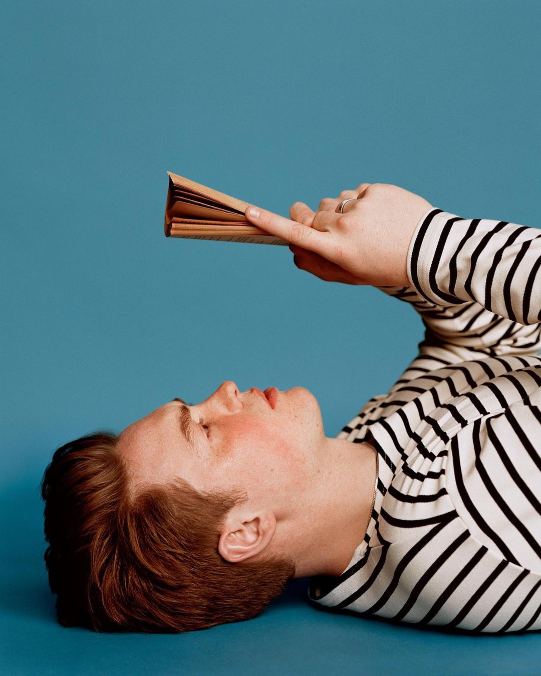 Adult male model lies on a blue background wearing a striped shirt, while holding a book open and pointing his finger at the pages.