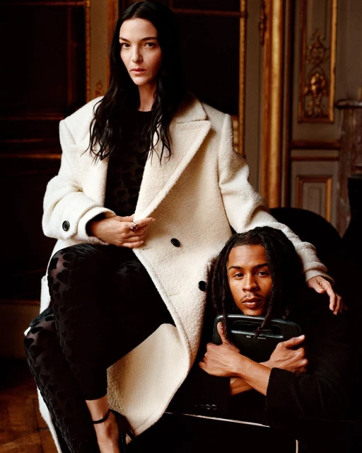 Elegant portrait of a woman in a cream coat and a man with dreadlocks holding a black bag, set against a backdrop of classic interior design.
