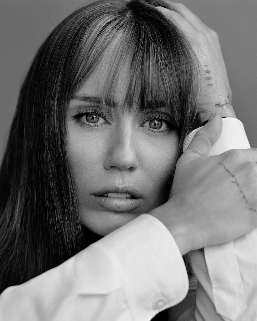 Monochrome portrait of an adult female with bangs and shoulder-length hair, wearing a white button-down shirt with hands near her face.