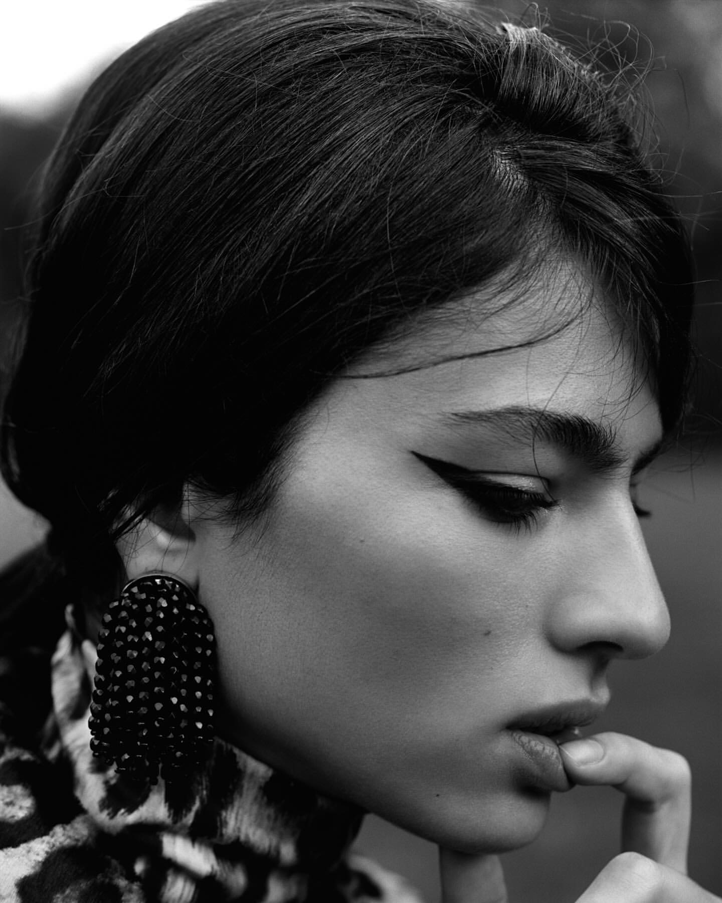 Monochrome portrait of an adult woman with dark hair and dramatic eye makeup, wearing large black fringe earrings and leopard-print top.