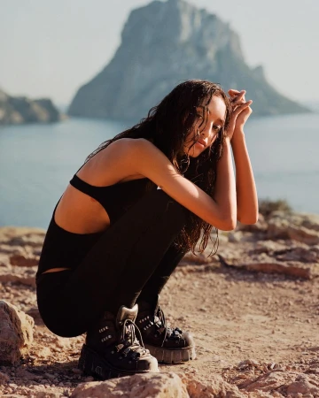 Woman with wet hair wearing black bodysuit and chunky boots crouching on a rocky coast before the ocean