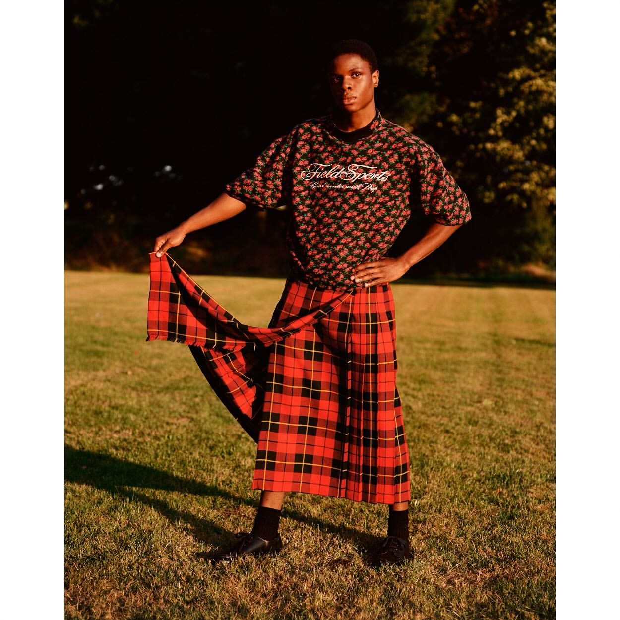Young adult poses outdoors on grass wearing a floral shirt and plaid kilt, creating a fashion-forward portrait with nature's backdrop.