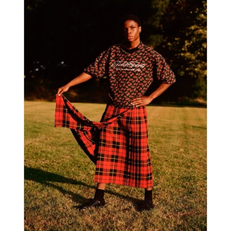 Young adult poses outdoors on grass wearing a floral shirt and plaid kilt, creating a fashion-forward portrait with nature's backdrop.
