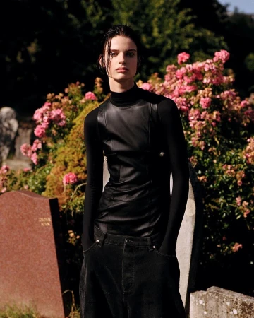A solitary, young adult model poses outdoors in a cemetery. The person wears a black leather vest over a long-sleeved black shirt and dark denim pants.