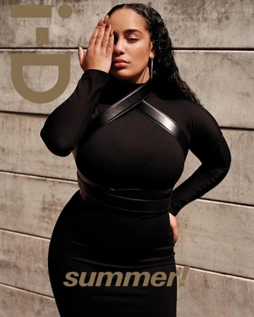 Woman in a black long-sleeve dress covering one eye with her hand against a concrete block wall