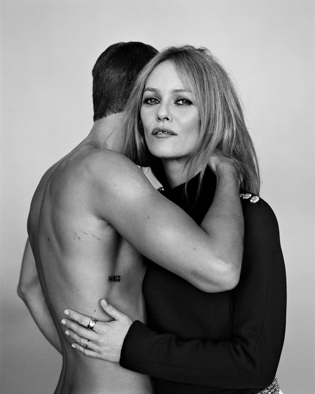 Black and white portrait of a blonde woman embracing a shirtless man from behind, against a plain background, showcasing a fashion editorial style.