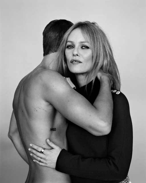 Black and white portrait of a blonde woman embracing a shirtless man from behind, against a plain background, showcasing a fashion editorial style.