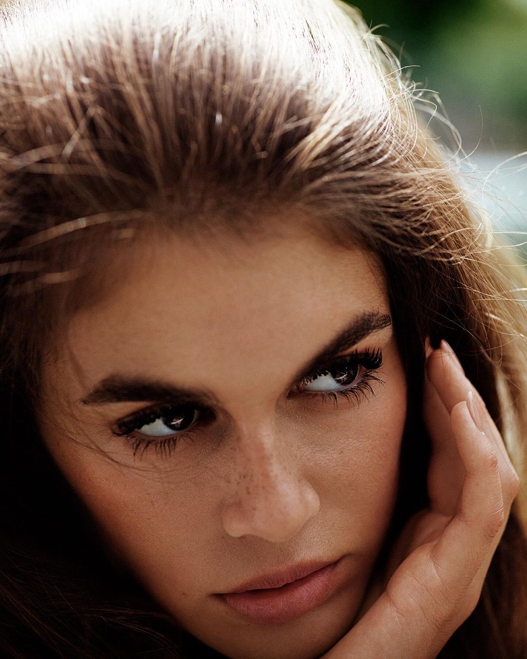 Close-up portrait of a woman with long brown hair touching her face, looking sideways with a serious expression.