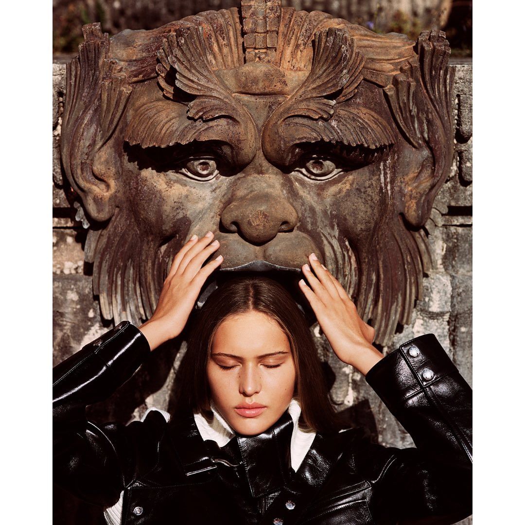 Young woman in black jacket poses beneath vintage stone mask outdoors, eyes closed, hands raised to her head, soft natural light.