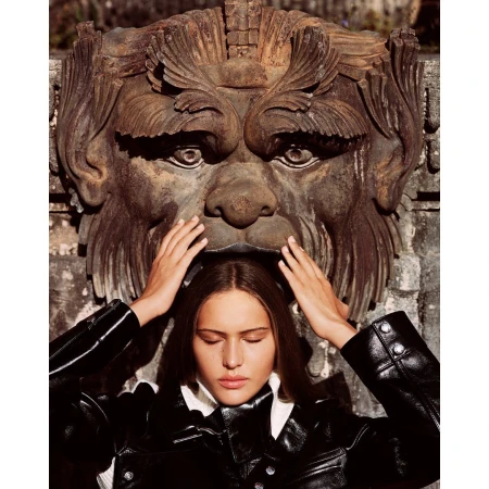 Young woman in black jacket poses beneath vintage stone mask outdoors, eyes closed, hands raised to her head, soft natural light.