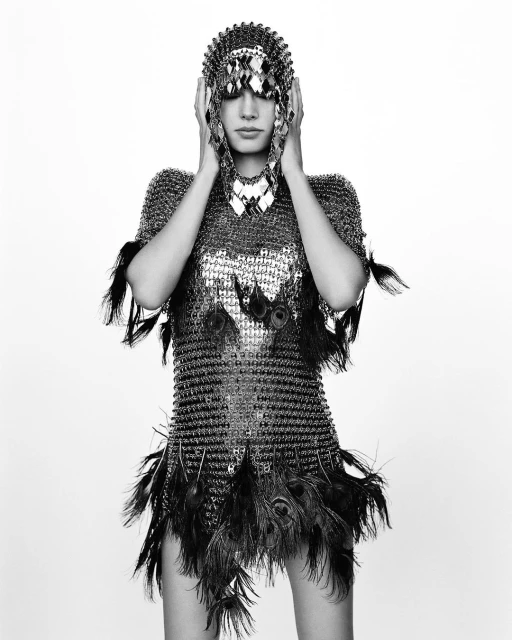 A woman in a metallic chainmail dress with peacock feathers posing in a studio setting, wearing a matching headdress in black and white.