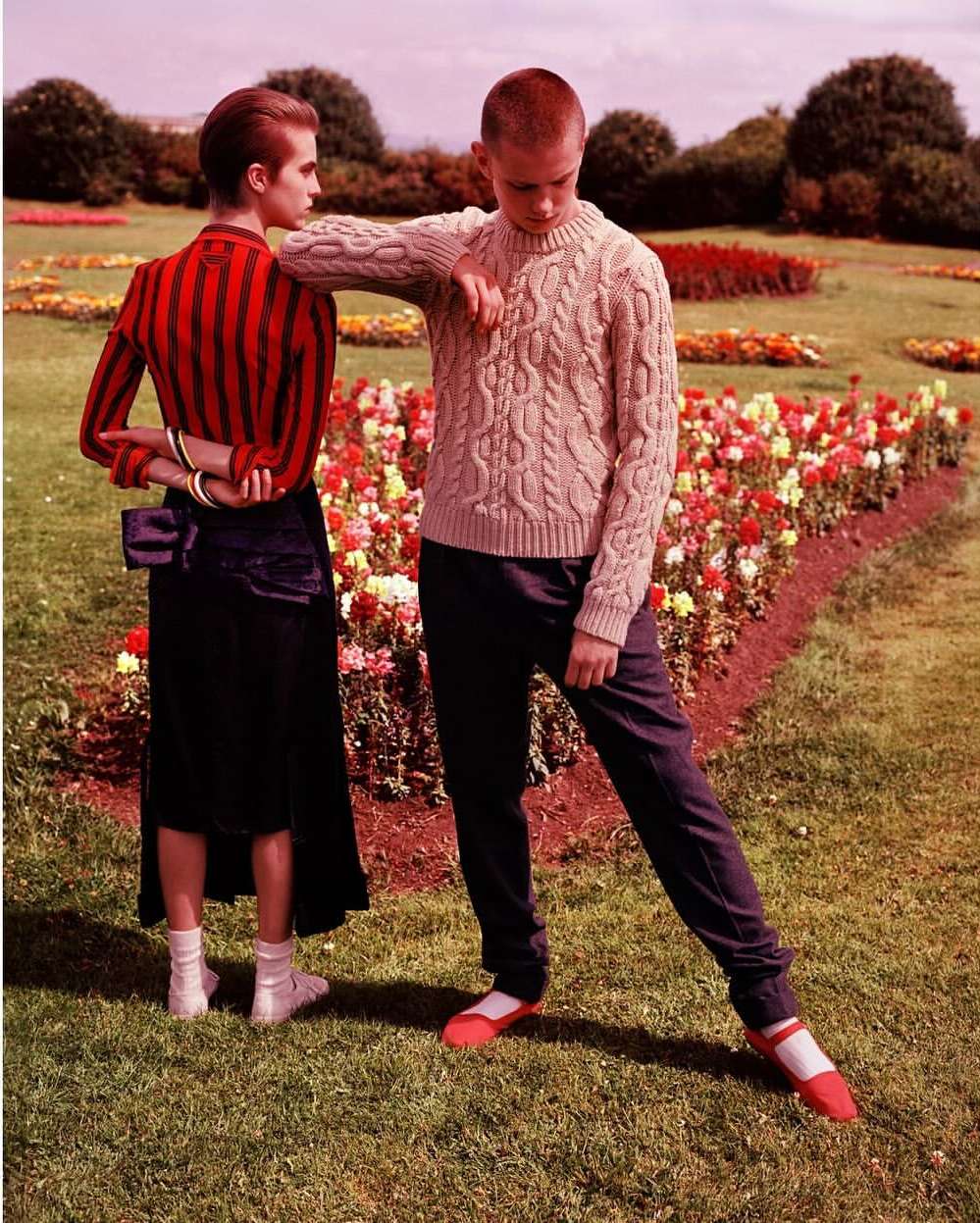 Two people pose in a garden with flower beds and green grass. The person on the left wears a red striped shirt and long black skirt.