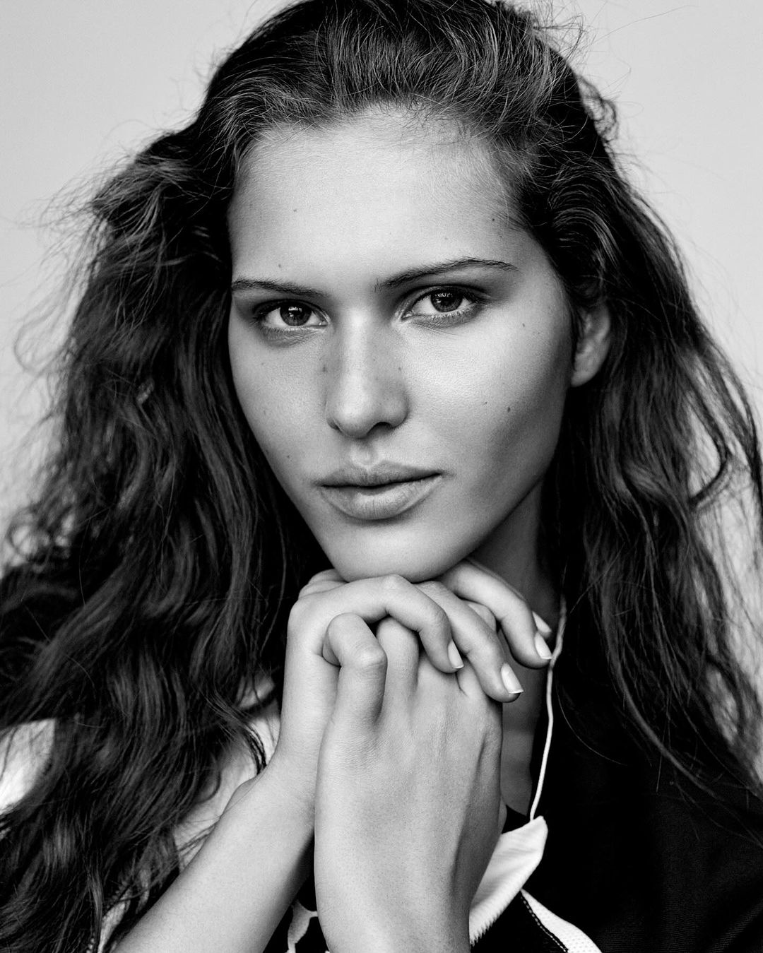 Black and white portrait of a woman with long wavy hair, her hands clasped under her chin, looking directly at the camera.