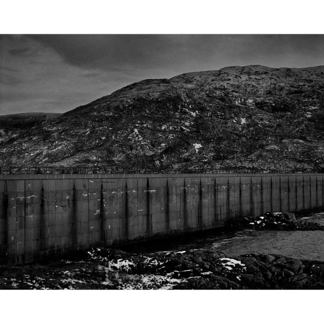 Monochrome landscape featuring a large concrete dam in the foreground and a hill covered with sparse vegetation in the background under a cloudy sky.