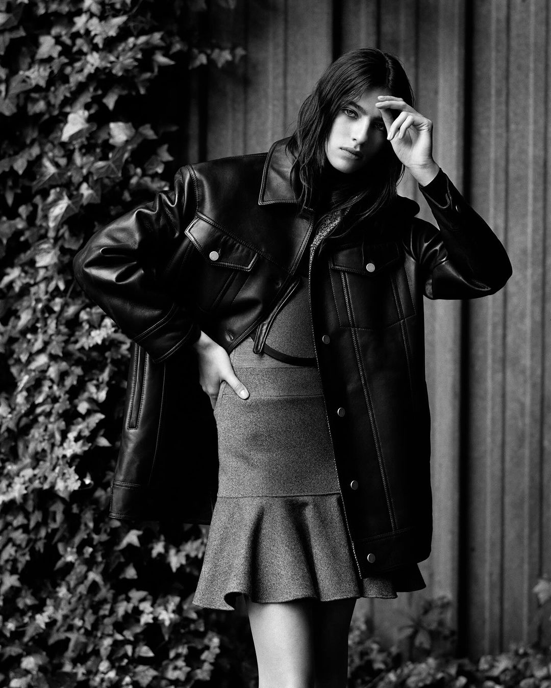 Monochrome fashion shot of an adult woman with long dark hair posing outdoors in a leather jacket and mini skirt, with green vine background.