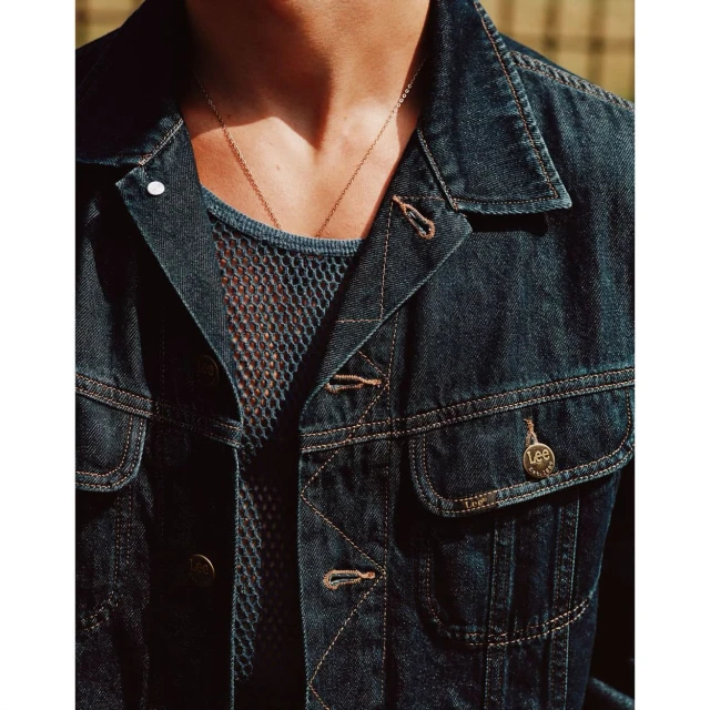 Close-up of an adult wearing a blue denim jacket over a dark fishnet shirt with a gold chain necklace in a brightly lit outdoor setting.