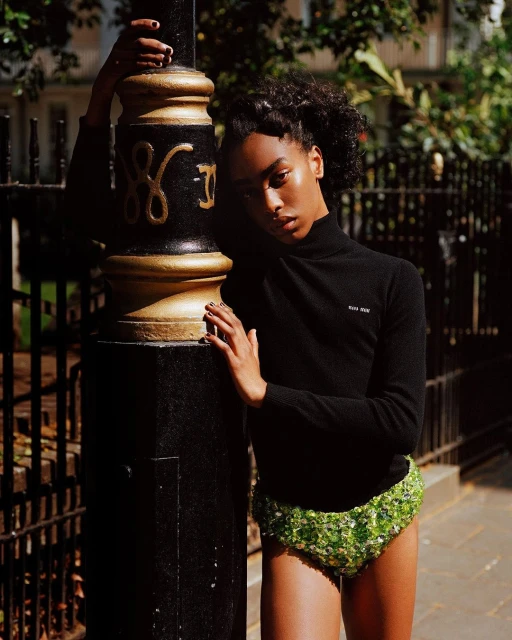 Woman poses in black top and green sequin shorts, leaning on a black and gold bollard, outdoors in dappled sunlight.