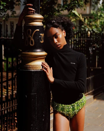 Woman poses in black top and green sequin shorts, leaning on a black and gold bollard, outdoors in dappled sunlight.