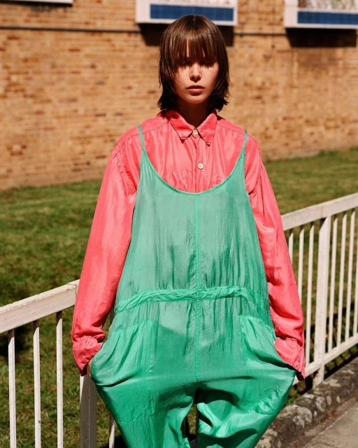 A young adult poses in a bright turquoise jumpsuit over a coral button-down shirt, standing against a brick wall and green lawn.