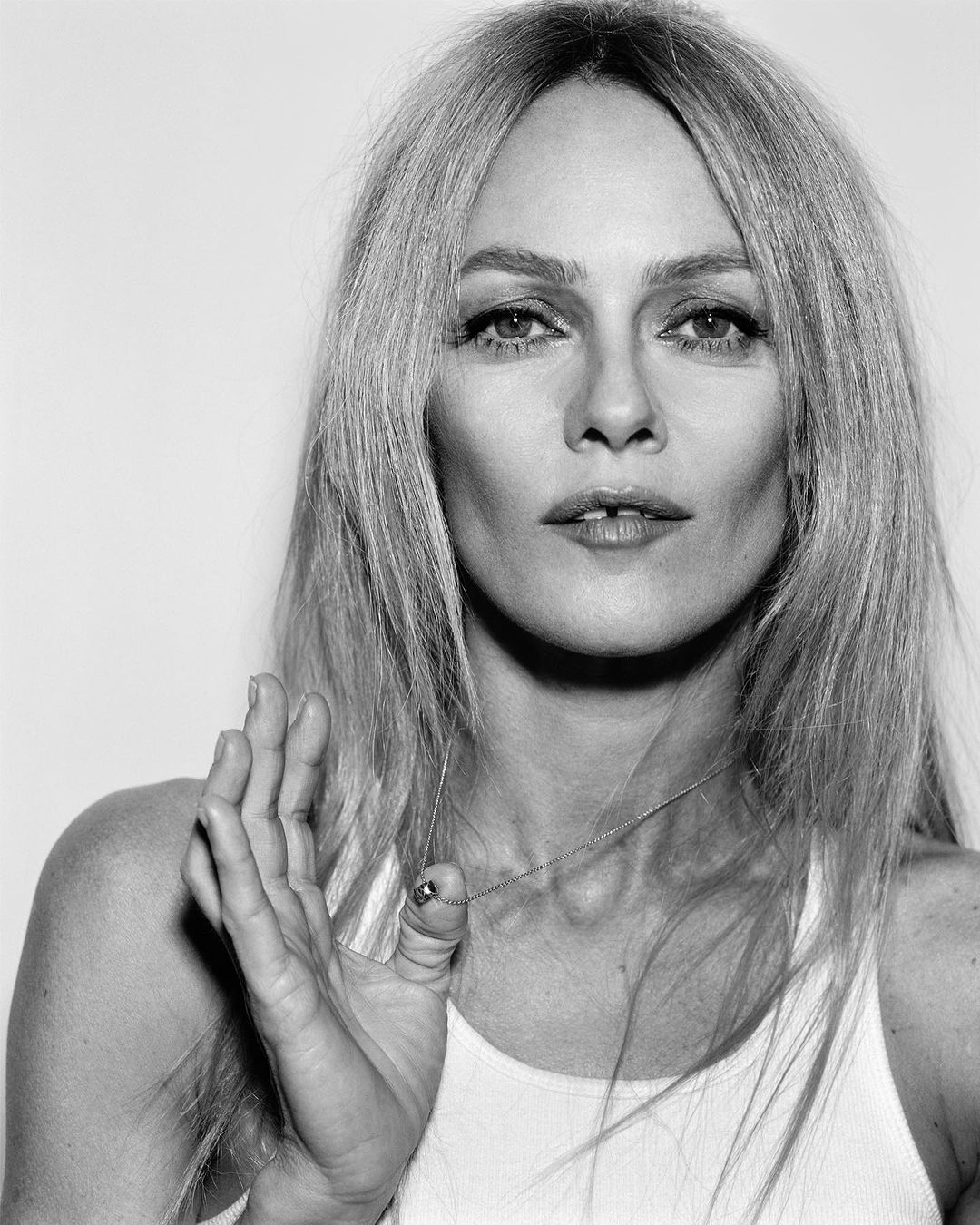 Close-up portrait of a blonde woman wearing a white tank top and a delicate pendant necklace against a plain backdrop.