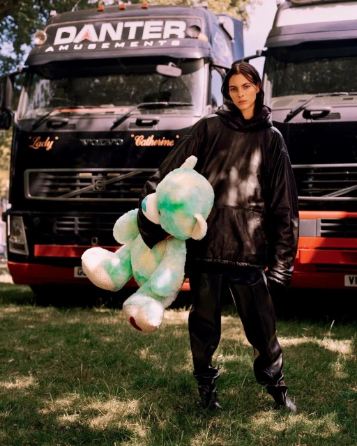 Adult female in a black leather outfit holding a stuffed bear in front of two black Danter Amusements trucks in a grassy outdoor setting.