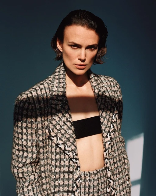 Keira Knightley poses in a tweed blazer with black crop top and a blue studio backdrop, highlighted by shadows and direct sunlight.