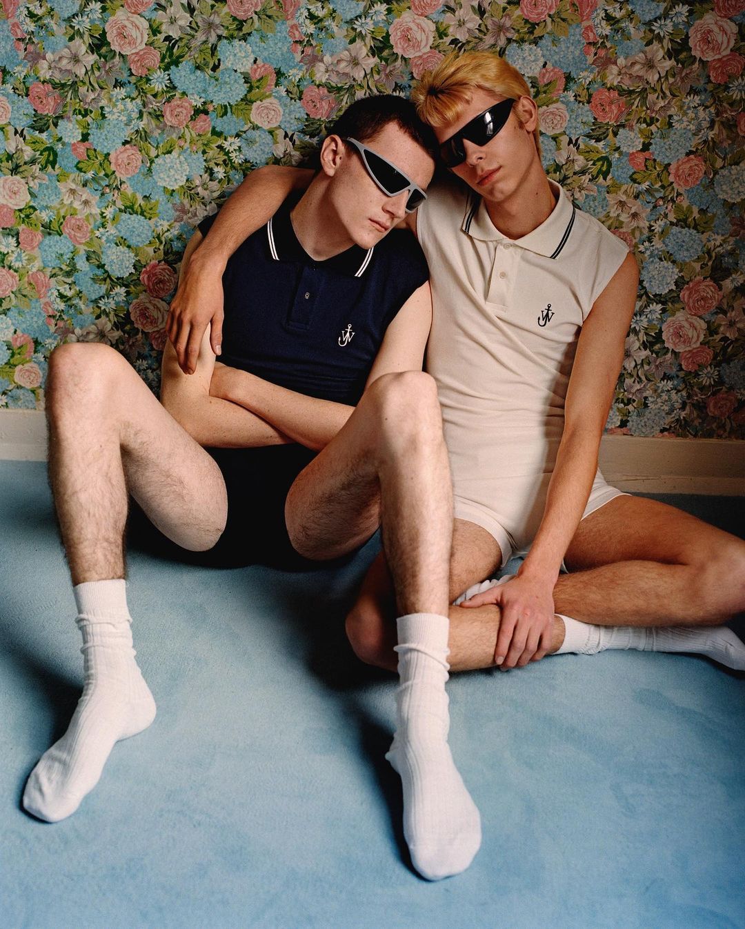Two men pose together in a polo shirt and underwear, seated on the floor against a floral wallpaper backdrop, showcasing a fashion-forward aesthetic.
