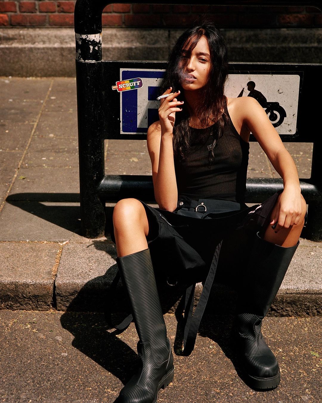 A woman in a black tank top and skirt sits on a curb smoking a cigarette in an urban environment with a sign in the background.
