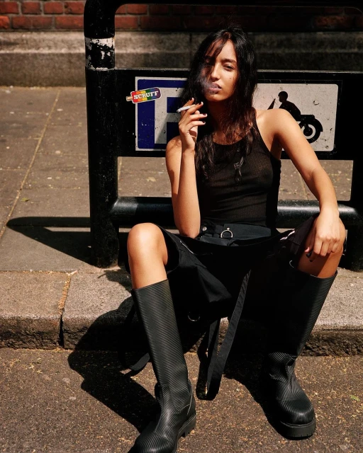 A woman in a black tank top and skirt sits on a curb smoking a cigarette in an urban environment with a sign in the background.