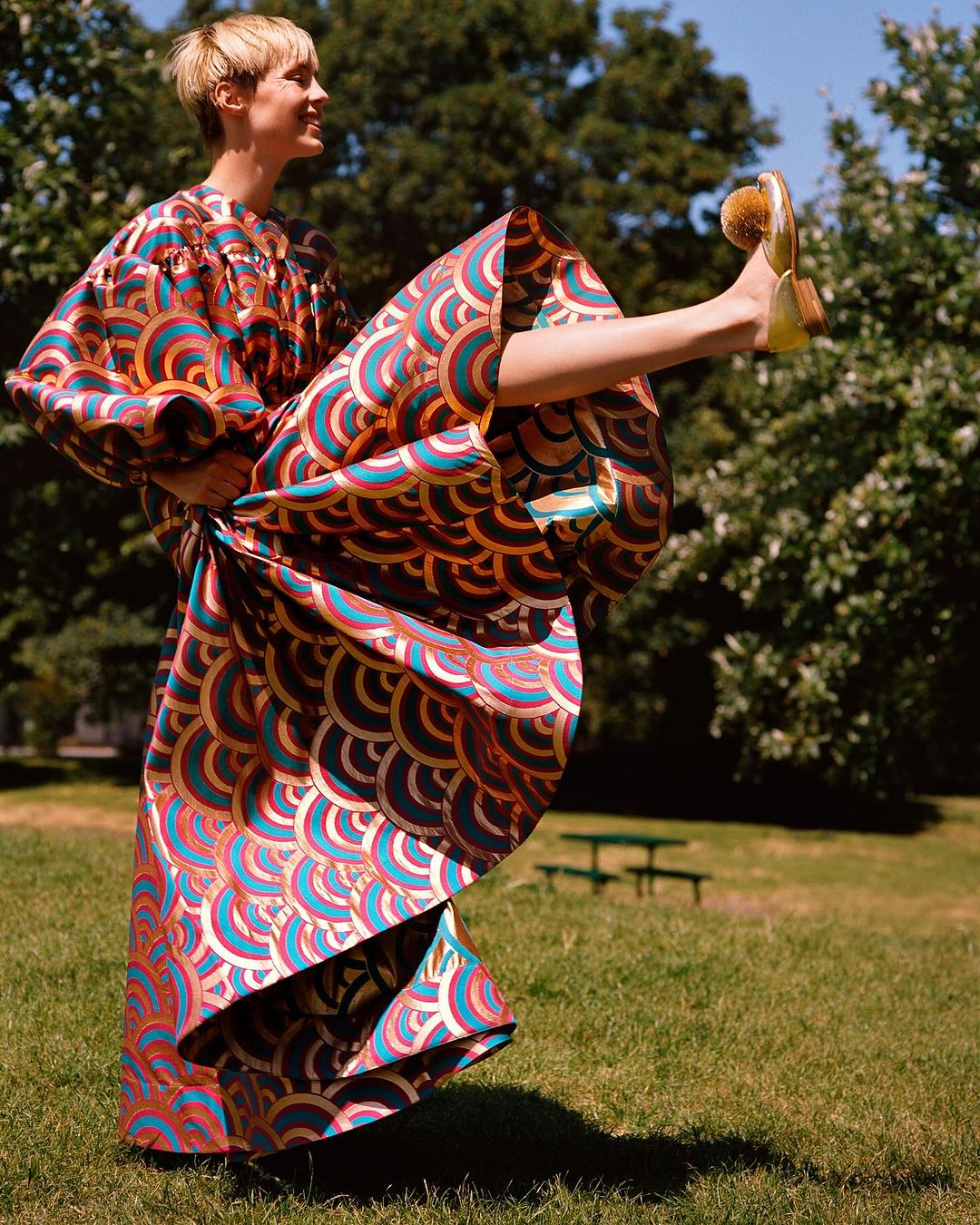 Woman in patterned dress with orange and blue, striking a playful pose with one leg raised outdoors against a backdrop of green trees.