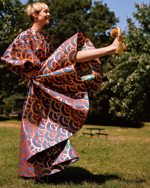 Woman in patterned dress with orange and blue, striking a playful pose with one leg raised outdoors against a backdrop of green trees.