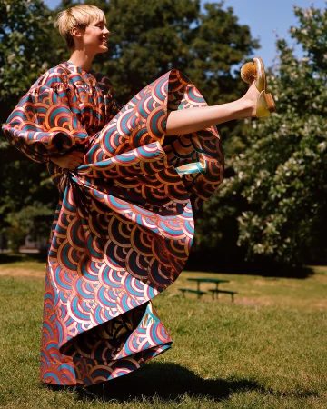 Woman in patterned dress with orange and blue, striking a playful pose with one leg raised outdoors against a backdrop of green trees.