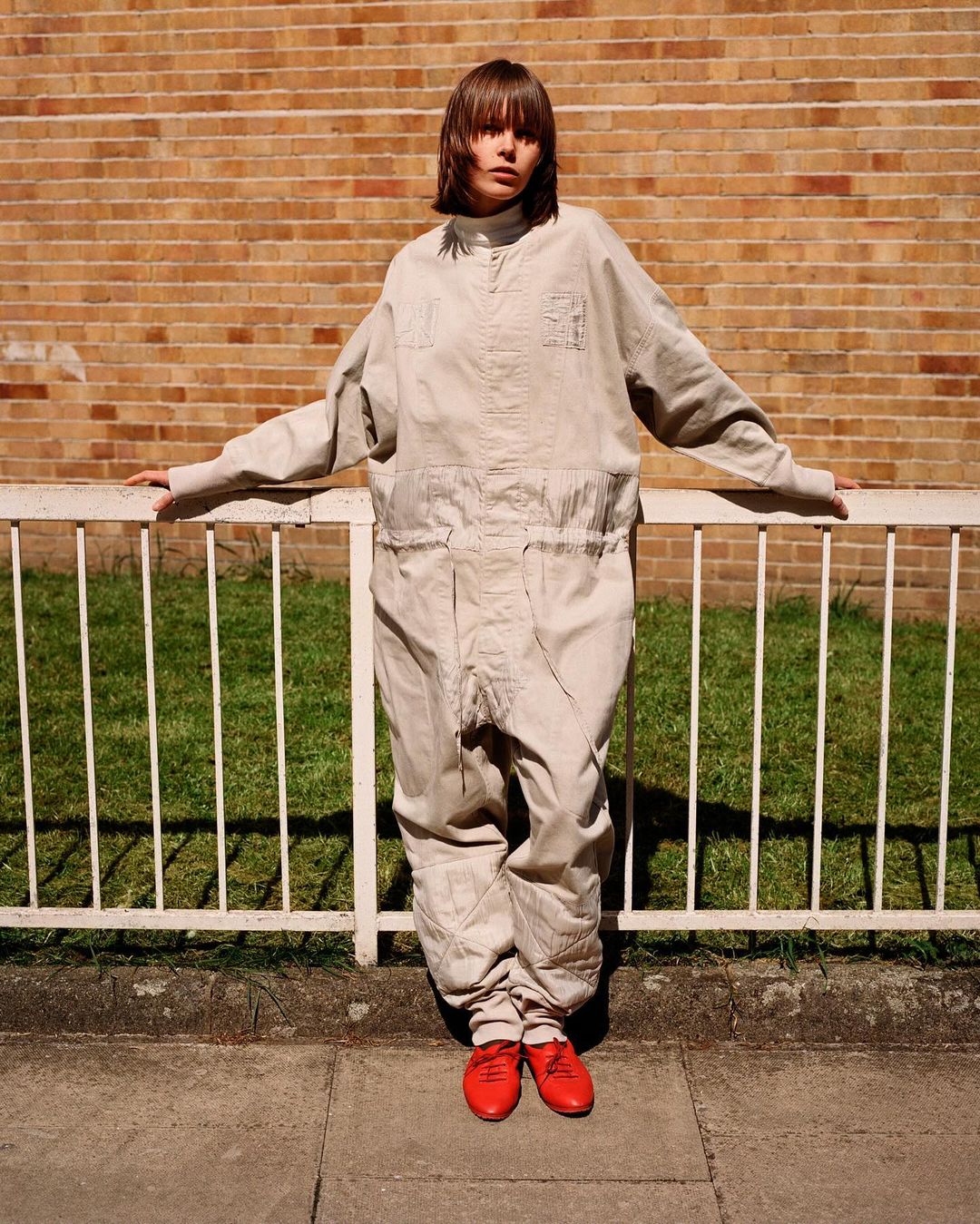 Model in cream jumpsuit leaning against white metal railing in front of a brick wall, wearing red shoes