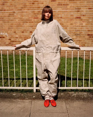 Model in cream jumpsuit leaning against white metal railing in front of a brick wall, wearing red shoes