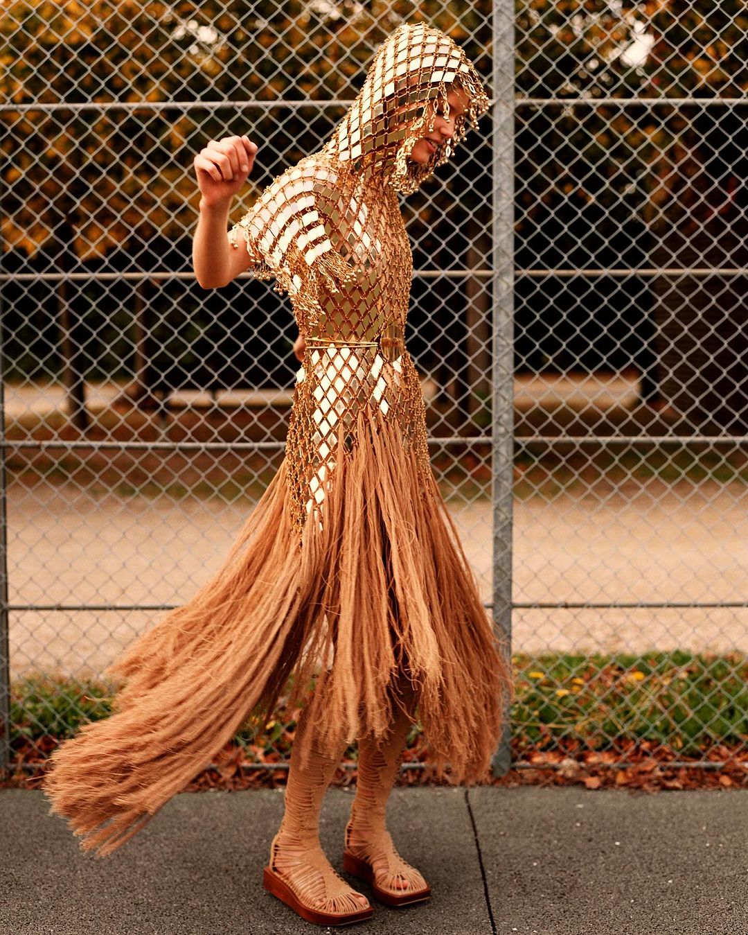 Adult female model wears gold fringe dress and hooded headpiece, posing on an outdoor sports court with chainlink fence in background.