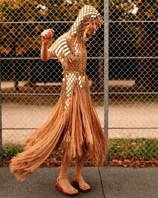 Adult female model wears gold fringe dress and hooded headpiece, posing on an outdoor sports court with chainlink fence in background.