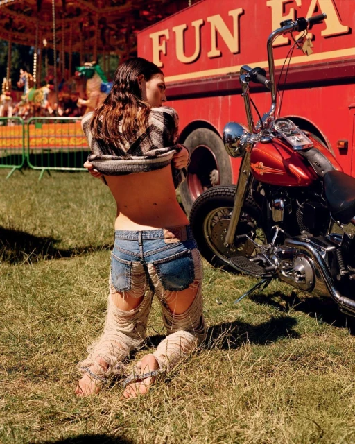 A woman kneels in ripped jeans, pulling up her shirt near a motorcycle and carnival tent on a grassy field in daylight.