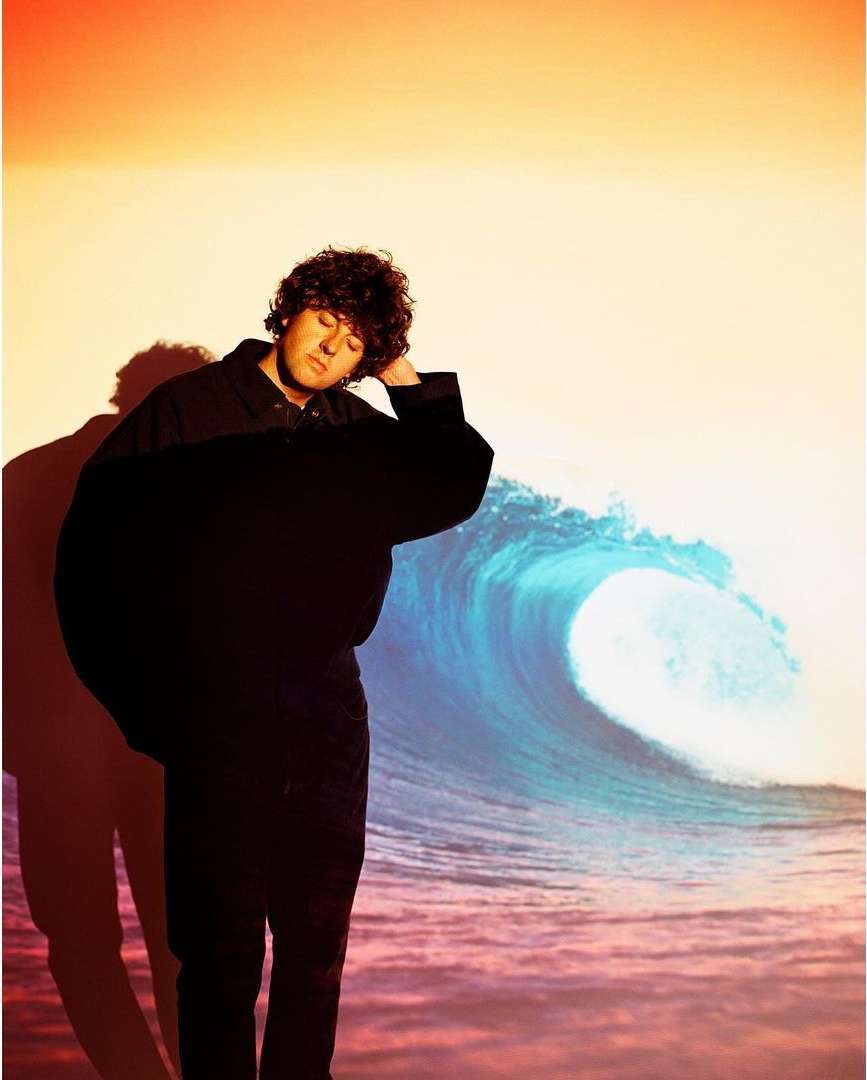 A man with curly hair wearing a dark jacket standing against a projection of an ocean wave and sunset sky.