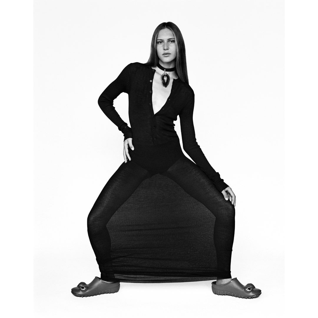 A long-haired model poses in a black dress and brown clogs against a white backdrop in a studio fashion shoot, with long shadows.