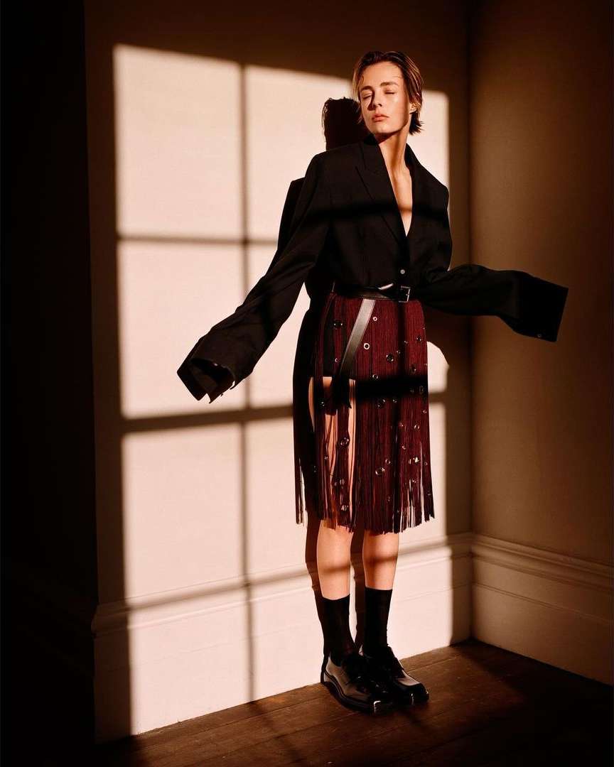 Adult female posing in a photo studio with dark jacket and fringed skirt, standing in window light with eyes closed in editorial fashion.