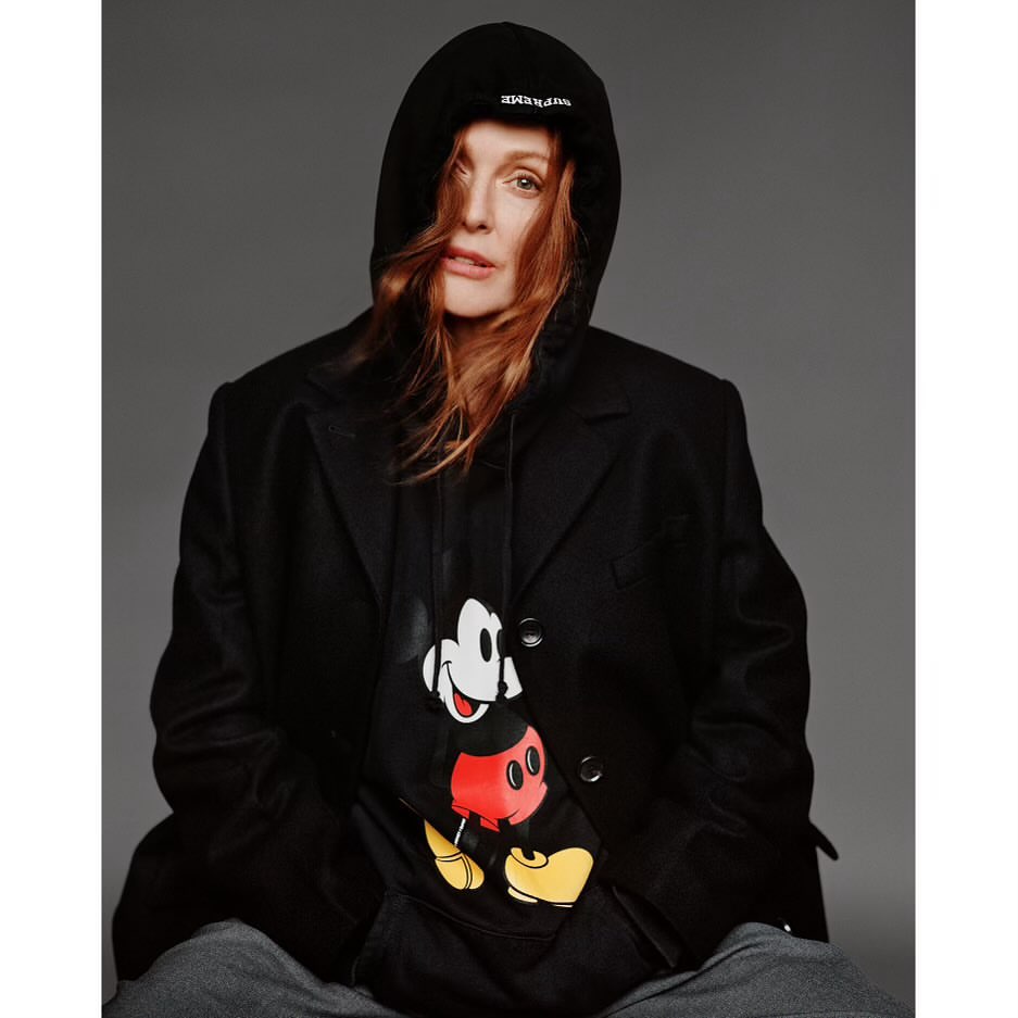 Adult woman with red hair wears a hooded sweatshirt with a Mickey Mouse graphic, under a dark coat, photographed in a studio.