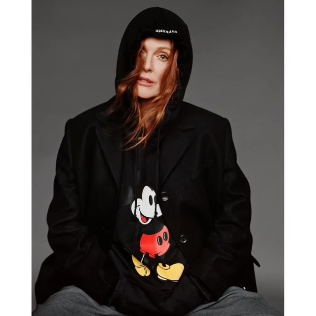 Adult woman with red hair wears a hooded sweatshirt with a Mickey Mouse graphic, under a dark coat, photographed in a studio.